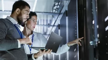 Two IT professionals work in a server room, using a laptop.