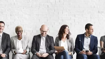 Group of professionally dressed people sitting against a white brick wall, smiling.