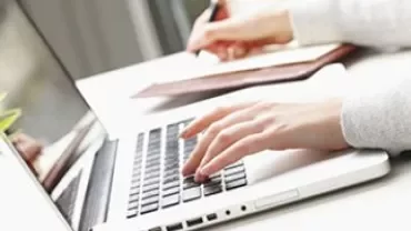 Person som skriver på en laptop och antecknar vid ett skrivbord.