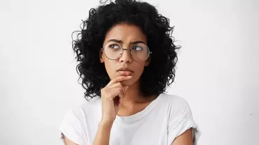 Person with curly hair thinking, wearing glasses and a white shirt.