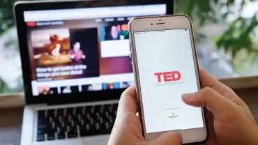 Person använder TED-appen på en smartphone med en bärbar dator i bakgrunden.