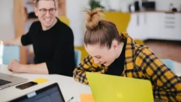 Two people laughing while working at laptops in an office.