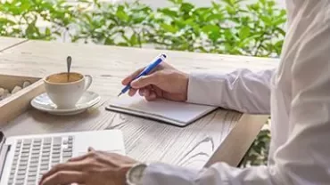 Person writing in notebook with laptop and coffee on desk.