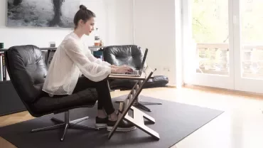 Person sitting on a leather chair using a laptop on an angled stand in a bright room.
