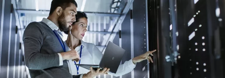 Two IT professionals work in a server room, using a laptop.