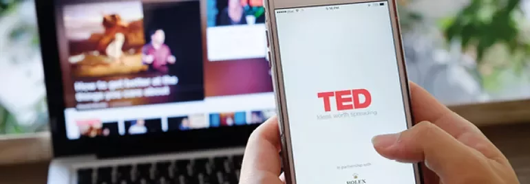 Person använder TED-appen på en smartphone med en bärbar dator i bakgrunden.