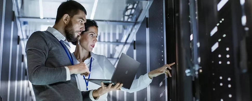Two IT professionals work in a server room, using a laptop.