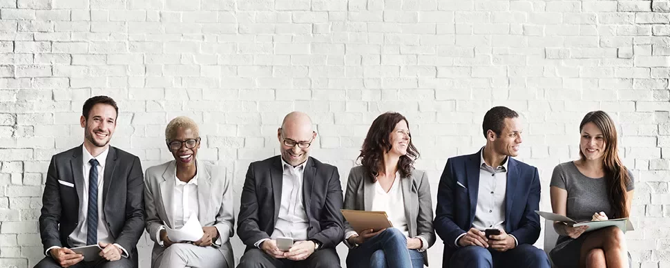 Group of professionally dressed people sitting against a white brick wall, smiling.